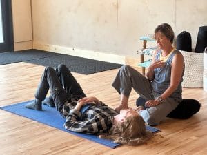 Physical therapy duluth mn. Robin Davidson sits near Kobi Jones discussing yoga and breathing. Kobi is laying on a yoga mat. the floor is natural hardwood. The wall is an off white lime paint.