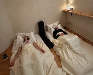 2 people lay on futons in a white plaster room with eye pillows over their eyes. They are covered in blankets and have bolsters near them.