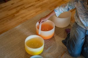 Sound mediation practitioner Kimberlee Grogan plays one of 3 large crystal sound bowls on a mat. The bowls colors fade from white to orange and yellow.